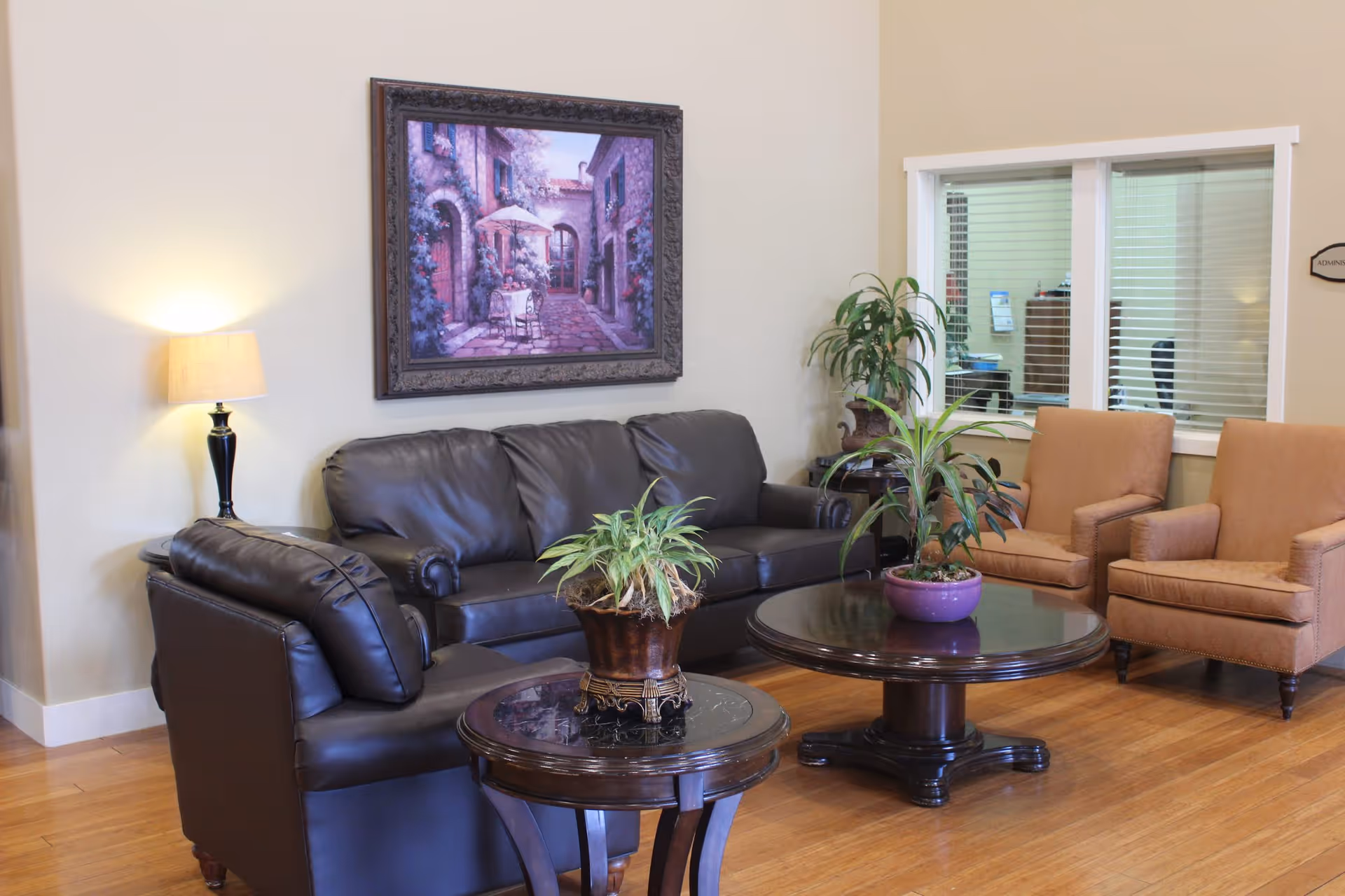 A cozy sitting area with a black leather sofa, two brown armchairs, and two round wooden tables with potted plants. A lamp is on a side table next to the sofa, and a framed painting of a quaint outdoor scene hangs on the wall. There is a window showing an office area in the background.