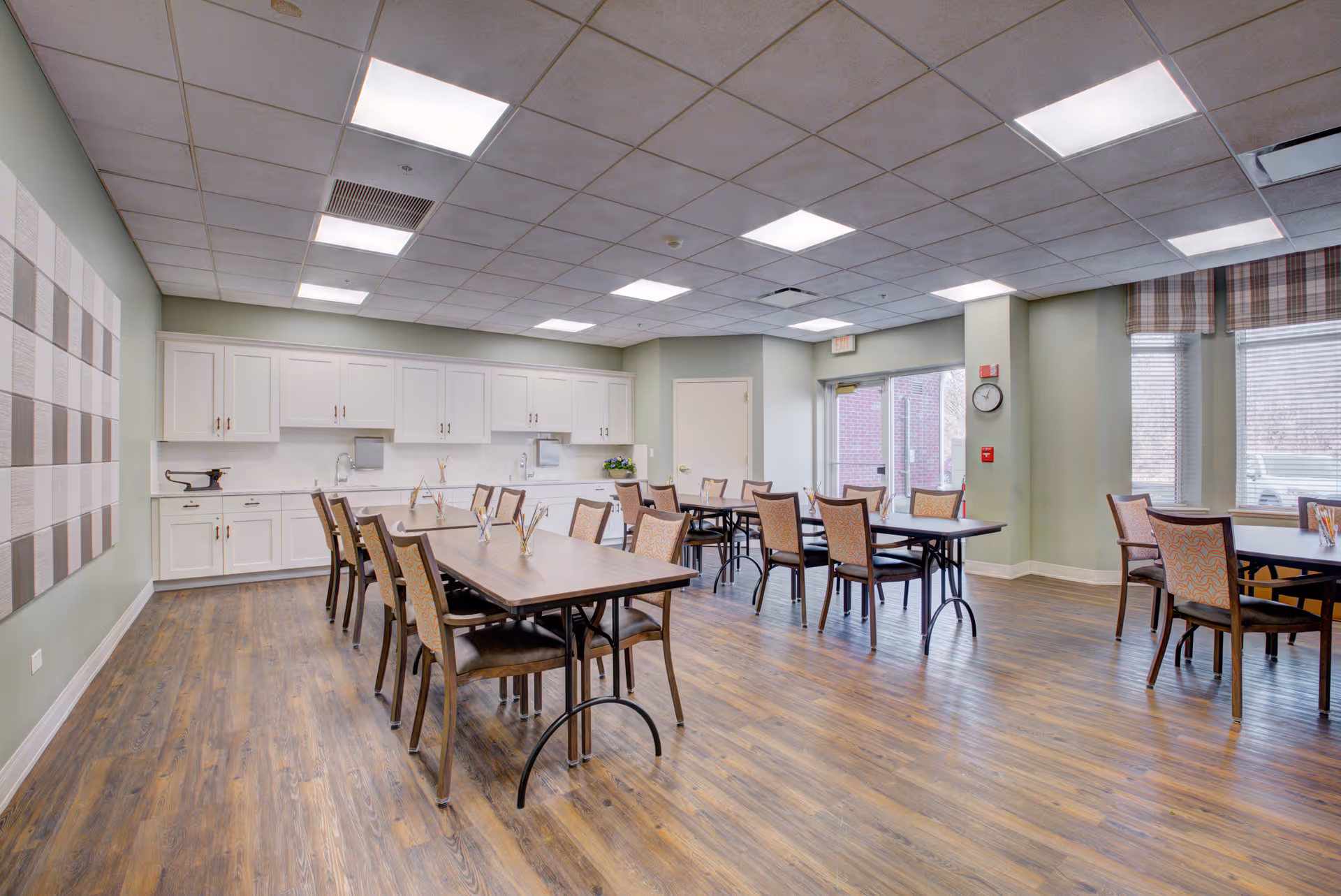 A bright dining room with several tables and chairs arranged neatly on a wooden floor. The room has light green walls, a large window with plaid valances, white cabinets along one wall, and ceiling lights providing ample illumination.