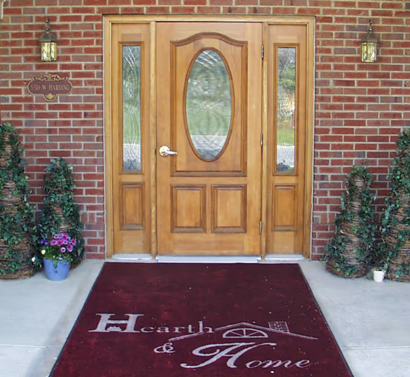 Front entrance of a building with a wooden door featuring an oval glass panel and two narrow glass side panels. The entrance is framed by red brick walls with two wall-mounted lantern lights on either side. There are decorative plants in woven baskets and a purple flower pot on the left side. A maroon floor mat with the text 'Hearth & Home' and a house logo is placed in front of the door.