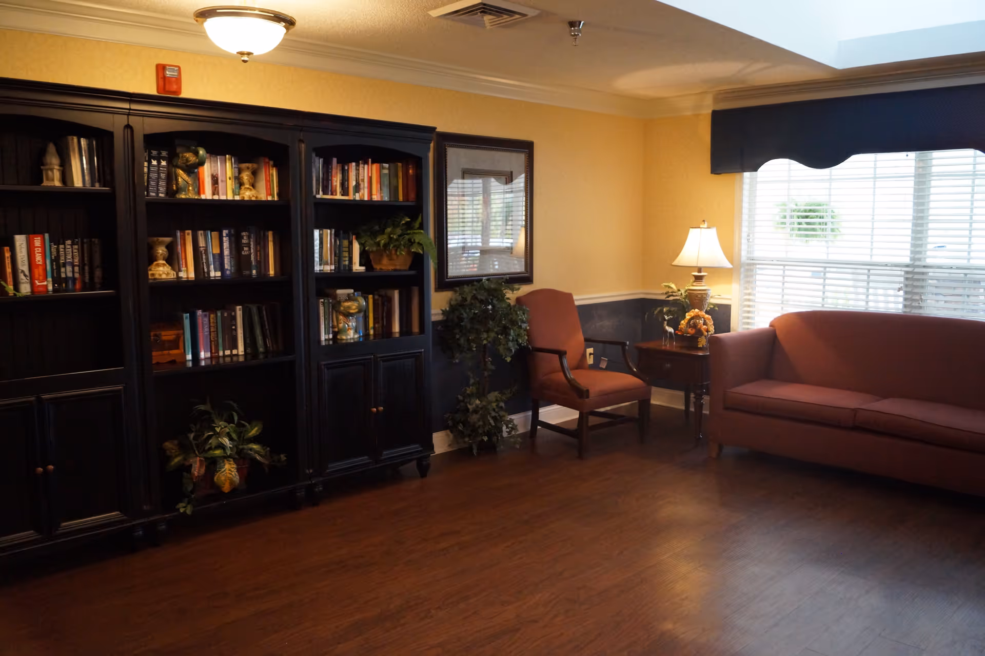 A cozy living room area with a dark wooden bookshelf filled with books and decorative items on the left, a framed picture on the wall, a potted plant, a maroon armchair, a side table with a lamp and floral arrangement, and a maroon sofa near a window with white blinds and a blue valance.