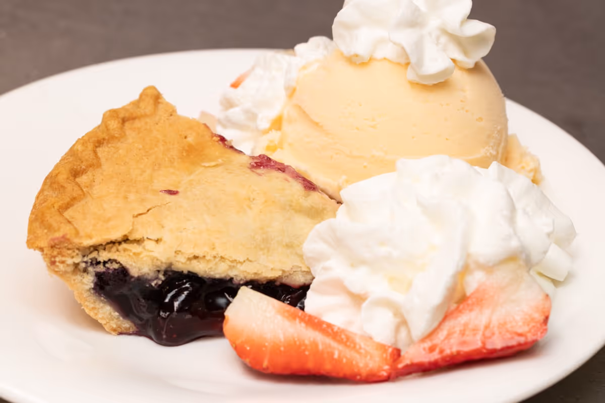 A dessert plate with a slice of blueberry pie, a scoop of vanilla ice cream topped with whipped cream, additional whipped cream on the side, and sliced strawberries.