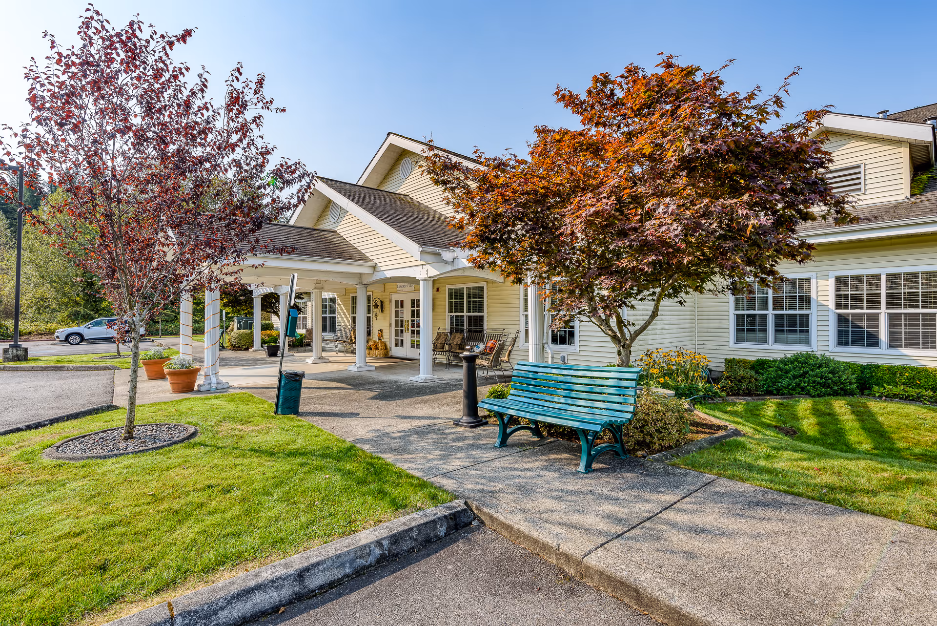 Exterior view of Cascade Place Assisted Living, Memory Care facility showing a beige building with white trim, a covered entrance with white pillars, a green bench under a tree with red leaves, and a well-maintained lawn and sidewalk.
