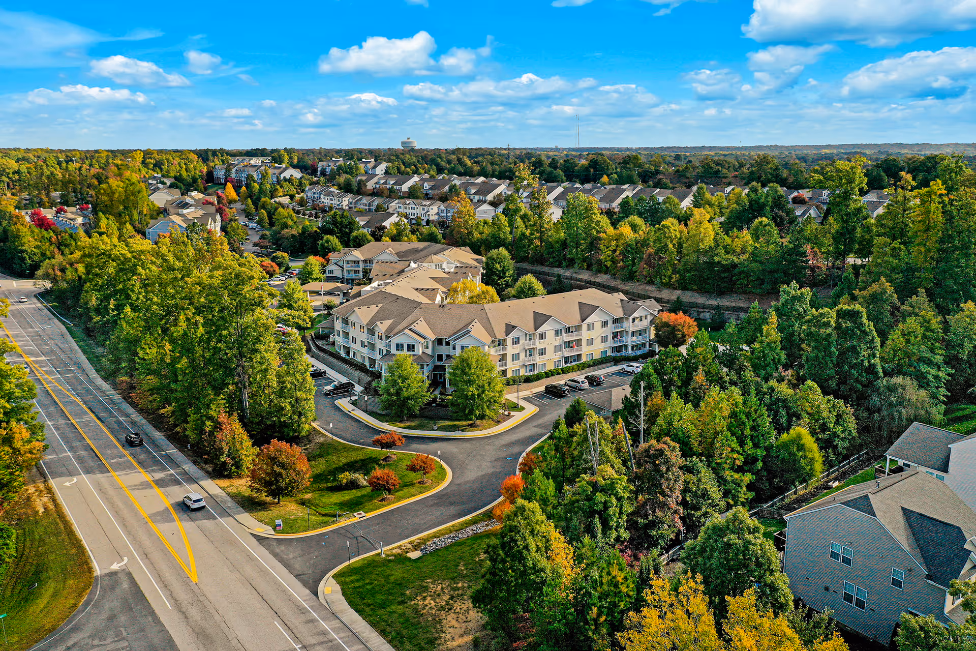Aerial view of Chesterfield Heights Gracious Retirement Living facility surrounded by trees and greenery, with a nearby road and residential neighborhood under a partly cloudy blue sky.