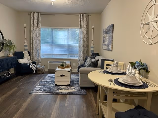 A cozy living room and dining area in an assisted living facility. The room features a gray sofa with patterned pillows, a small white coffee table on a blue and white rug, a navy blue armchair with a white throw, and a round dining table set for two with plates, bowls, and napkins. The walls are beige with a large decorative compass and a framed picture. A large window with patterned curtains lets in natural light, and the floor is dark wood.