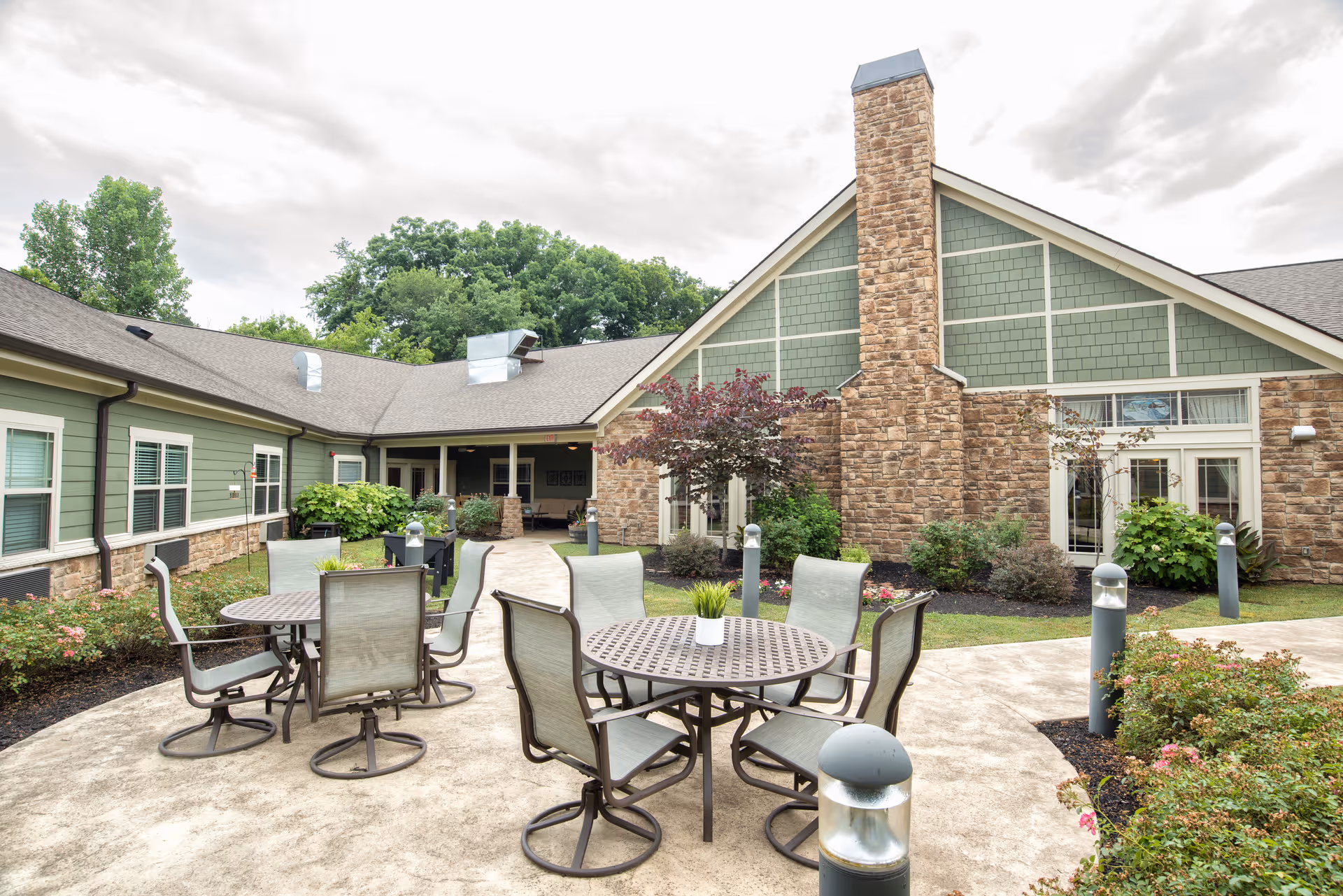 Outdoor patio area at Dominion Senior Living of Sevierville featuring round metal tables with chairs arranged around them, surrounded by landscaped greenery and a building with green siding and stone accents in the background under a cloudy sky.