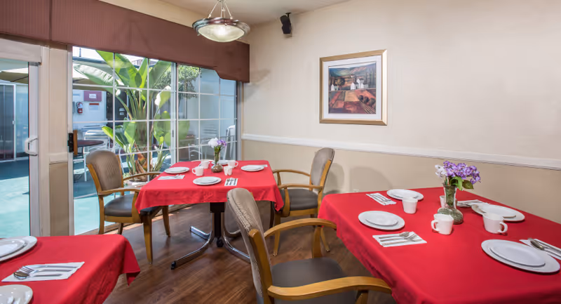 A dining room with three tables covered in red tablecloths, each set with white plates, cups, and silverware. There are wooden chairs with cushions around the tables. A large sliding glass door reveals an outdoor area with plants. A framed painting hangs on the beige wall, and a ceiling light fixture is visible.