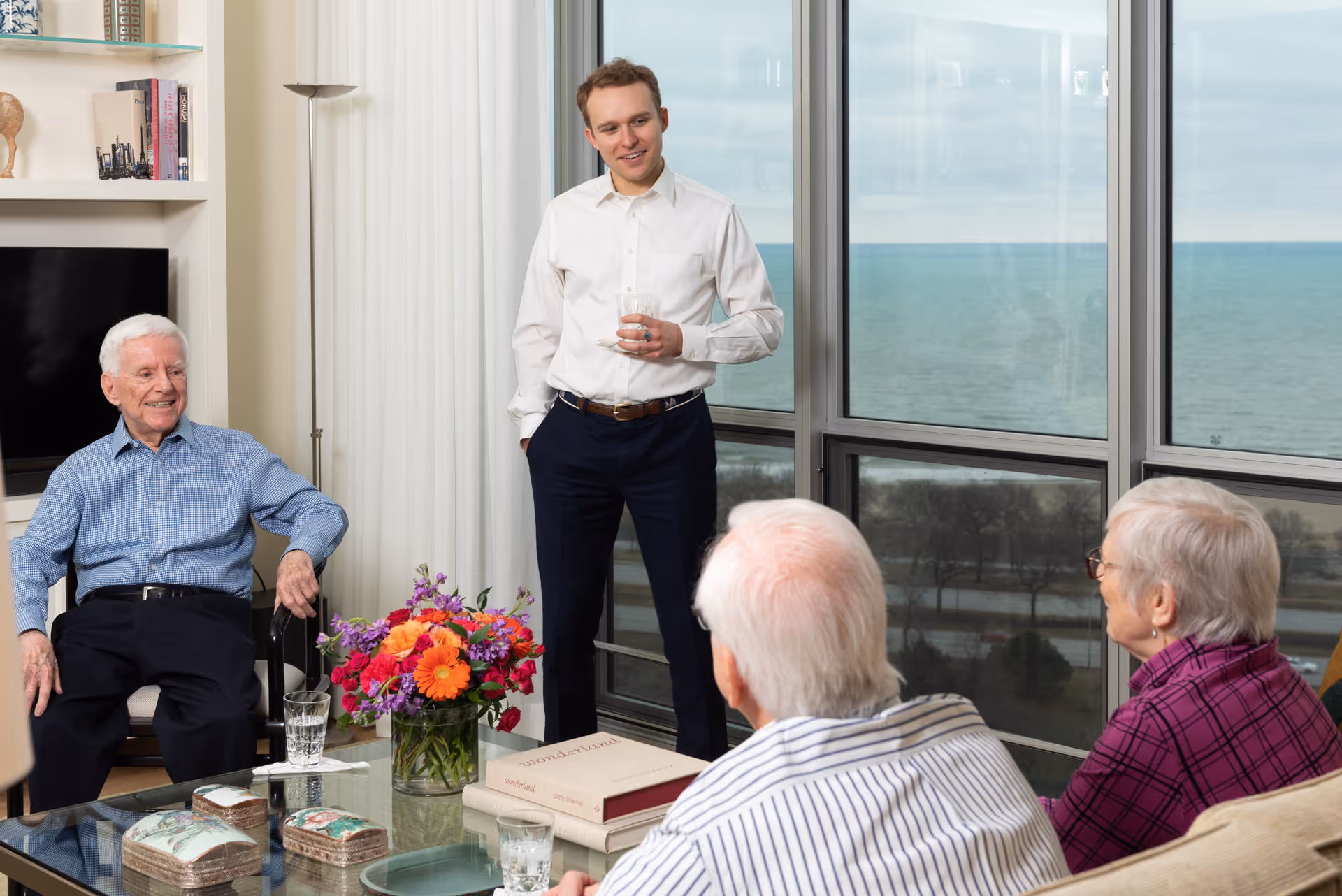 A group of elderly people and a younger man are gathered in a bright living room with large windows overlooking a body of water. The elderly man on the left is sitting and smiling, while the younger man stands holding a glass. Two elderly people are seated on a couch facing them. A glass coffee table with a colorful flower arrangement and books is in the center.