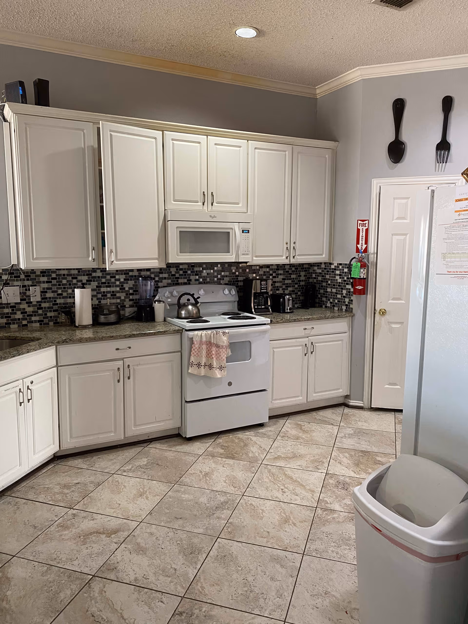 A clean kitchen with white cabinets, a white stove with a kettle on it, a microwave above the stove, a coffee maker, a toaster, and a blender on the countertop. The backsplash features a mosaic tile design in shades of gray and black. The floor is tiled with large beige tiles. A white trash can is visible in the foreground, and a fire extinguisher is mounted on the wall near a door. Decorative oversized wooden fork and spoon hang on the wall above the door.