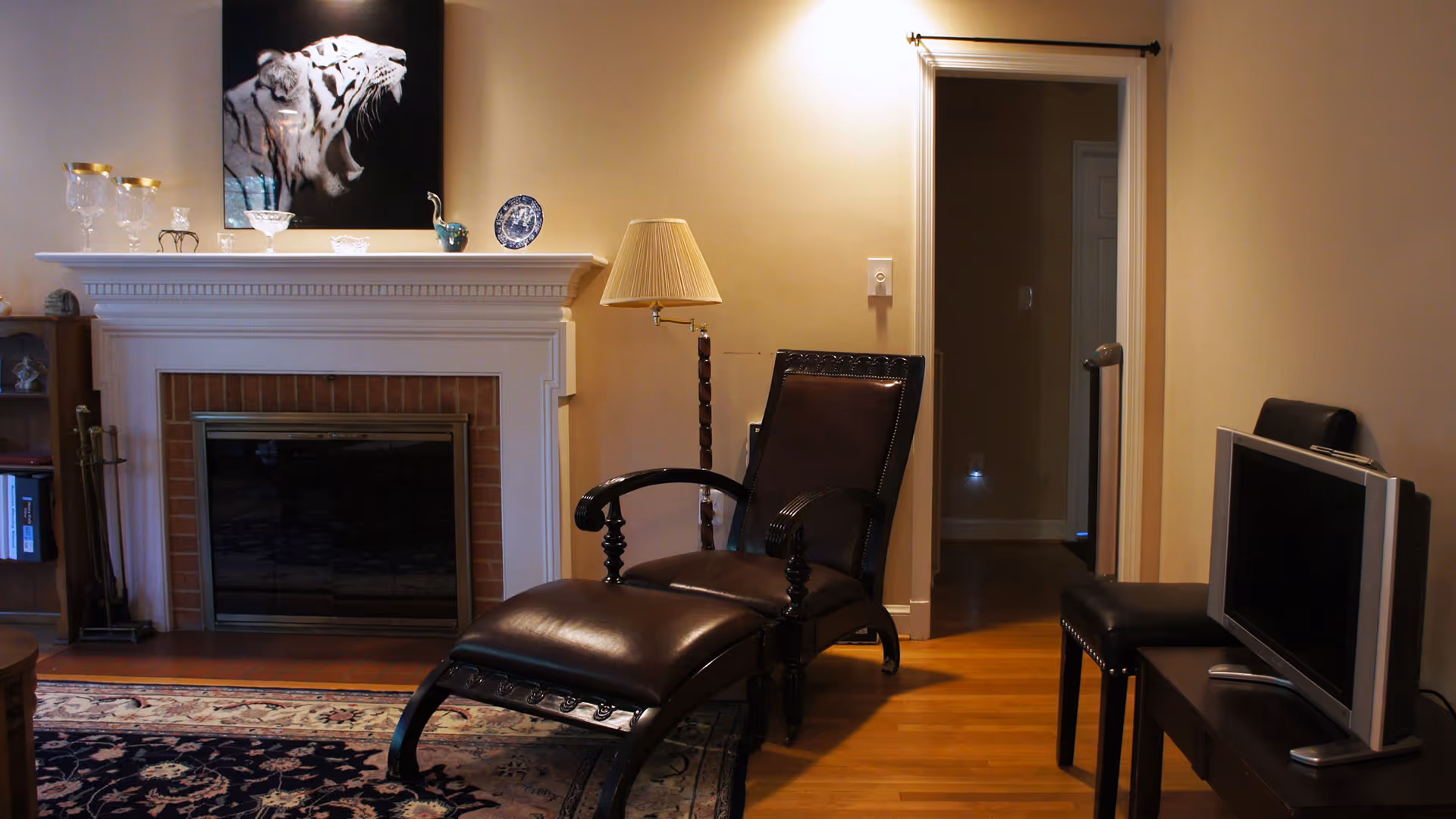 A cozy living room with a dark leather chaise lounge chair and a floor lamp next to a white fireplace mantel. The mantel is decorated with glassware and a decorative plate, and above it hangs a large black and white picture of a roaring tiger. To the right, there is a small TV on a stand with a chair beside it, and a doorway leading to another room.