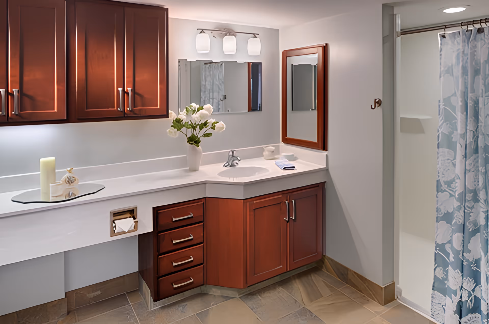 A clean and well-lit bathroom featuring a white countertop with a built-in sink, wooden cabinets and drawers with silver handles, a large mirror above the sink, a smaller wooden-framed mirror on the adjacent wall, a vase with white flowers on the counter, a candle and decorative items on a glass tray, and a shower area with a blue and white floral shower curtain.