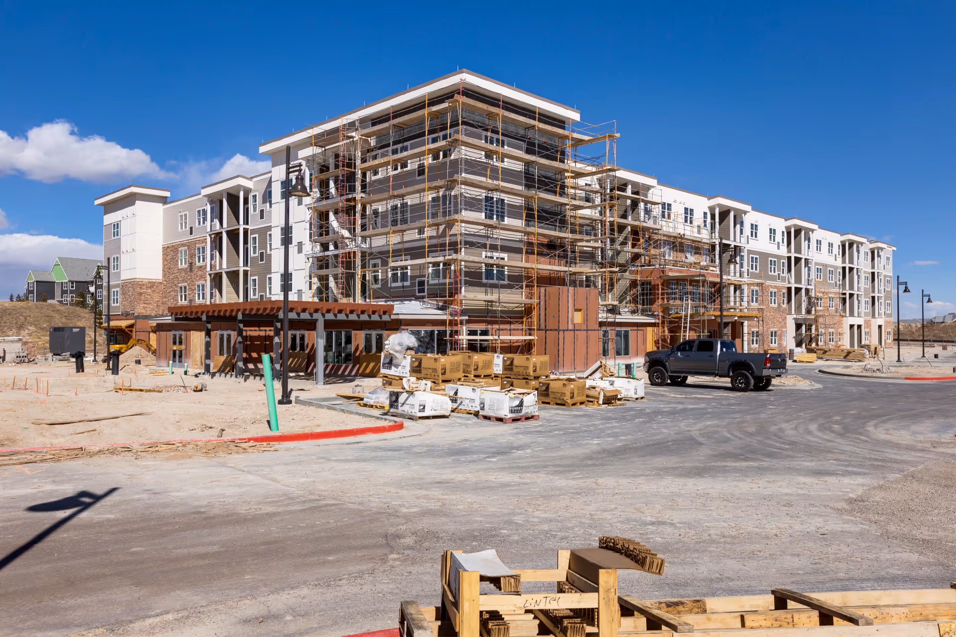 A large multi-story building under construction with scaffolding on the exterior. The building has a combination of brick and light-colored siding. Construction materials and equipment are scattered around the site, and a black pickup truck is parked near the building. The sky is clear with a few clouds.