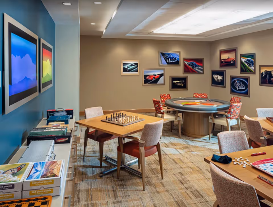 A cozy game room with several tables and chairs. One table has a chessboard set up, another has a bingo game in progress, and a round table in the back has a poker or card game setup. The walls are decorated with framed pictures of classic cars. Shelves along one wall hold board games and puzzles.
