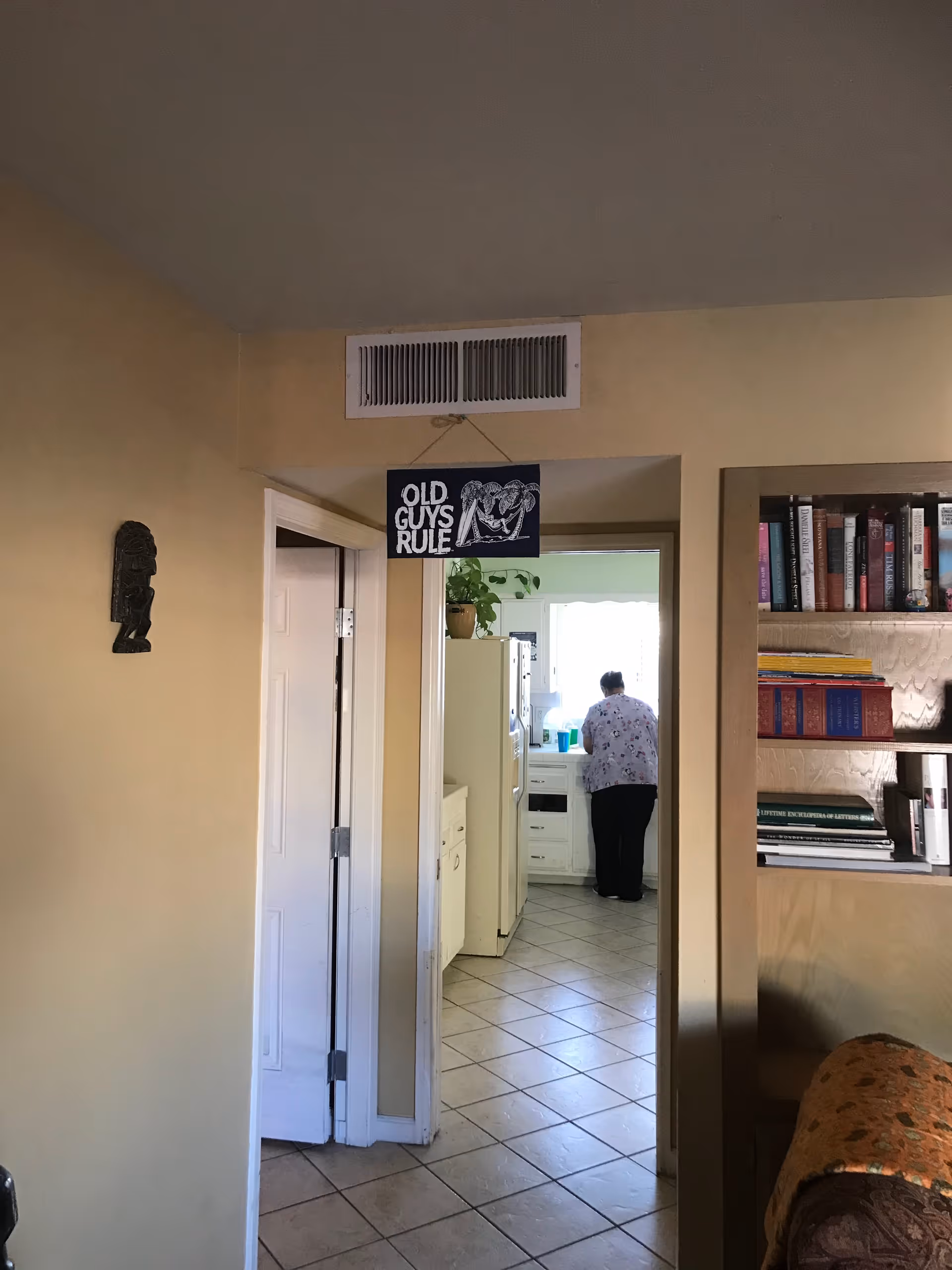View from a living room area into a kitchen where a person is standing at the counter. The walls are painted beige, and there is a sign hanging above the doorway that reads 'OLD GUYS RULE'. A bookshelf filled with books is visible on the right side, and a small decorative mask is mounted on the left wall.
