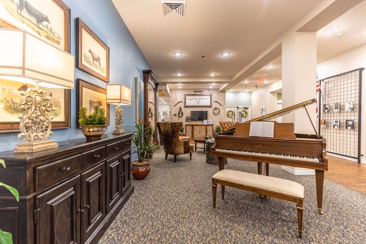 A warm assisted-living common room with a grand piano and bench, seating area, sideboard with lamps and framed artwork.
