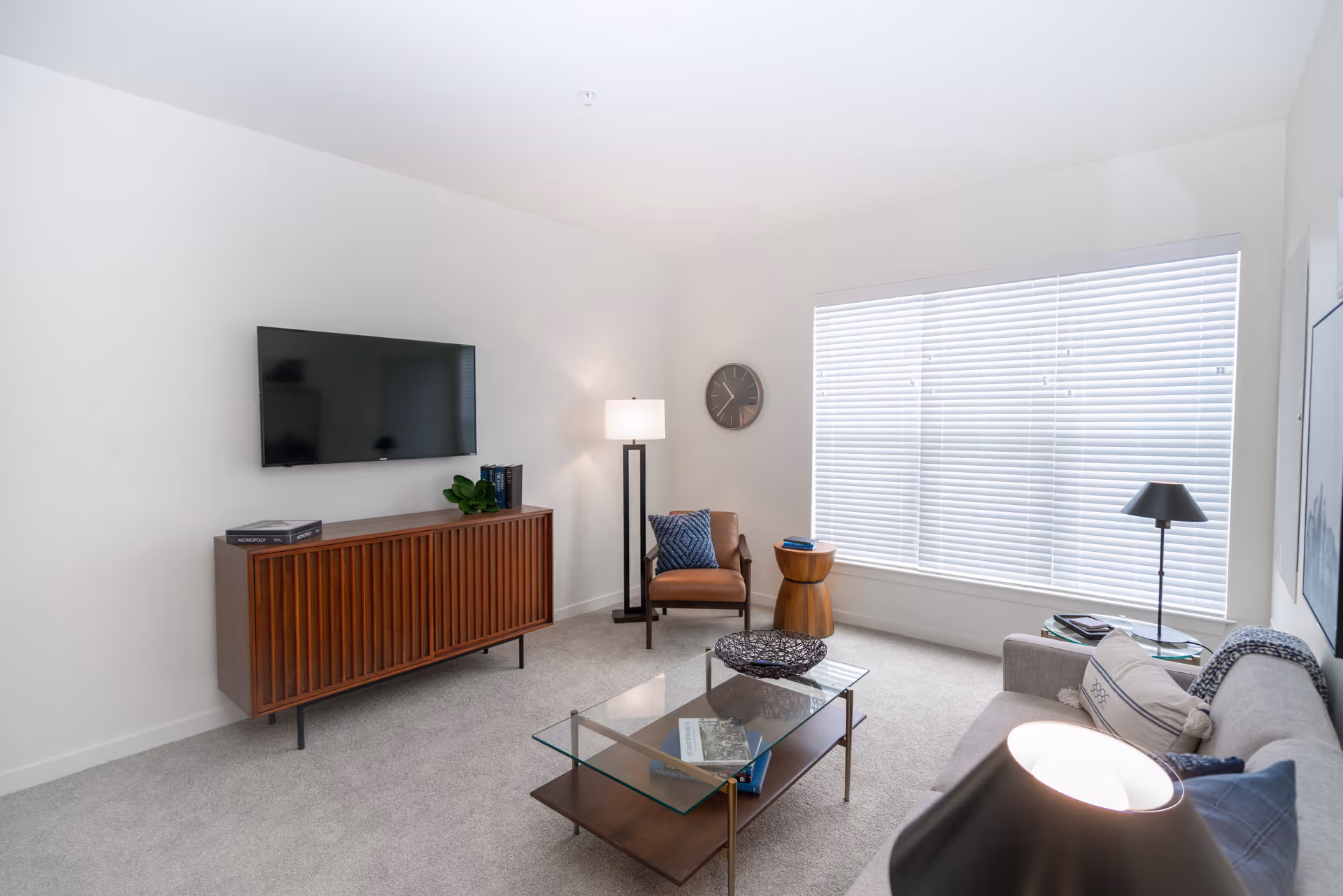 A bright living room with a large window covered by white blinds. The room features a wall-mounted flat-screen TV above a wooden cabinet, a brown leather armchair with a blue pillow, a floor lamp, a round wooden side table, a glass coffee table with books and decorative items, and a gray sofa with cushions. The walls are white and the floor is carpeted in light gray.