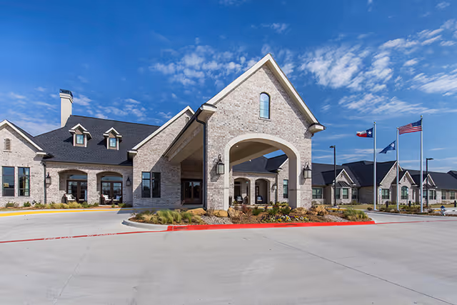 Exterior view of a senior living facility building with a large covered entrance, stone facade, multiple windows, and three flagpoles displaying the Texas state flag, the United States flag, and another flag. The sky is clear with some clouds.