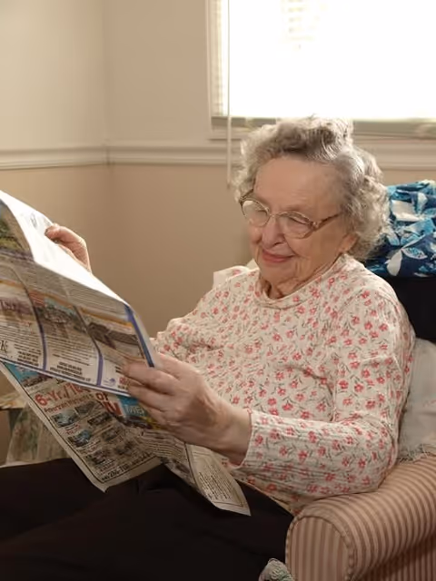 An elderly woman with glasses and curly gray hair is sitting comfortably in a striped armchair, reading a newspaper. She is wearing a light-colored floral blouse and appears content and relaxed in a well-lit room with a window behind her.