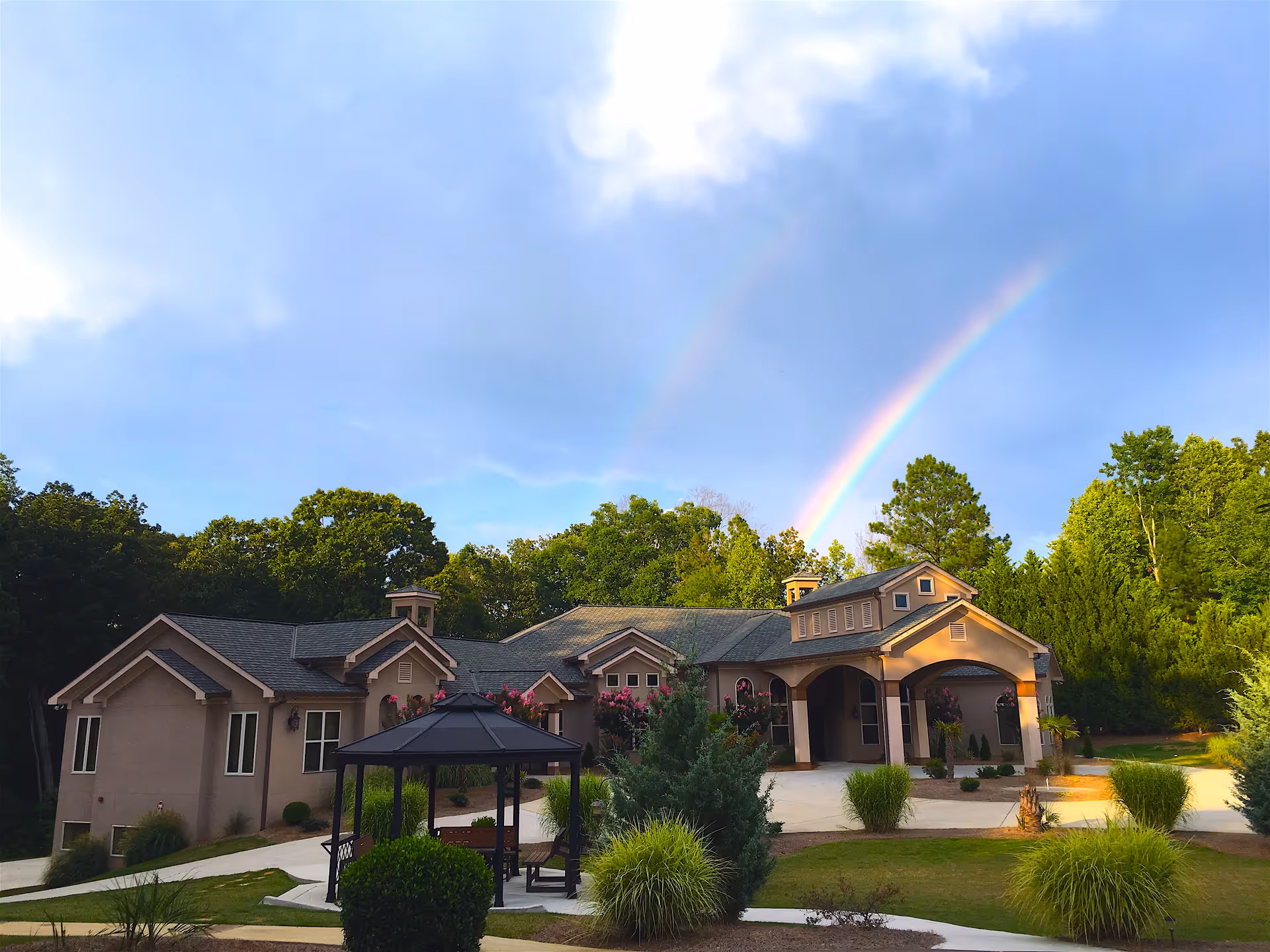 A single-story senior living facility building with a beige exterior surrounded by greenery and trees. In front of the building is a black gazebo with benches, and a well-maintained lawn with shrubs and plants. A vibrant double rainbow is visible in the sky above the building.