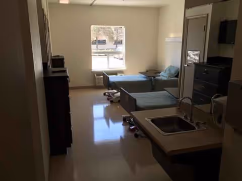 A compact hospital style patient room with bed, window, dresser, and kitchenette sink.