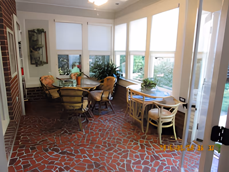 Sunroom seating area with wicker chairs and small tables on a brick-patterned floor beside large windows with shades.
