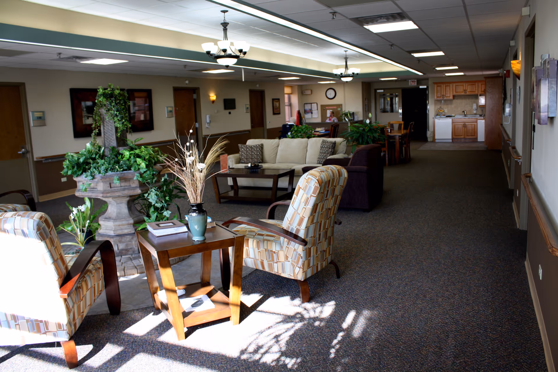 A well-lit common area in a senior living facility featuring two patterned armchairs around a small wooden table with a vase and books, a stone planter with green plants, a beige sofa with cushions, additional seating, and a kitchenette in the background.