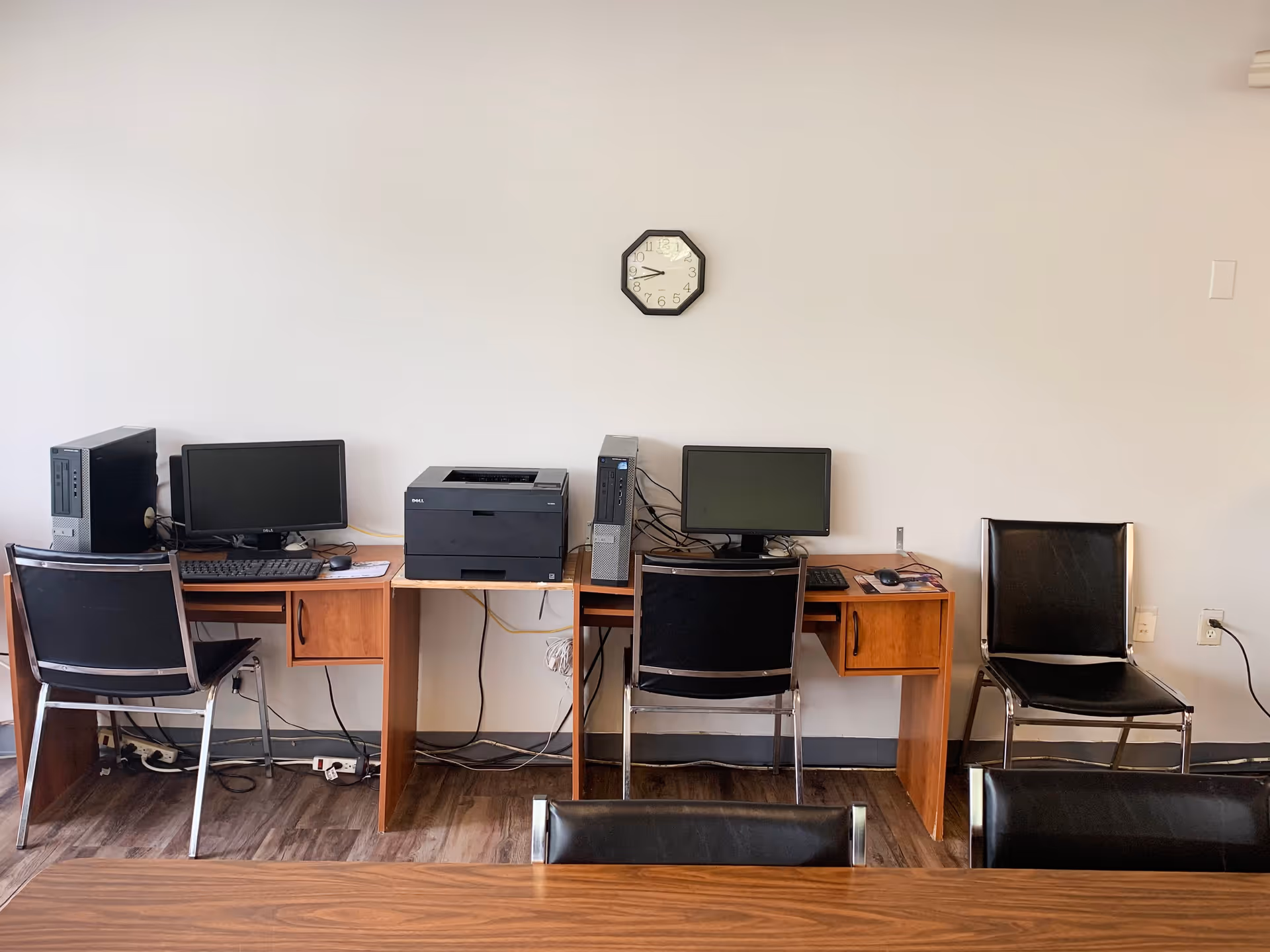 A small office area with two wooden desks against a plain white wall. Each desk has a black computer monitor and a desktop tower, with a black printer placed on a small table between the desks. Two black chairs with metal frames are positioned in front of the desks, and a third black chair is placed to the right. A simple octagonal wall clock is mounted above the desks, showing the time as 9:50. The floor is covered with wood-patterned vinyl flooring.