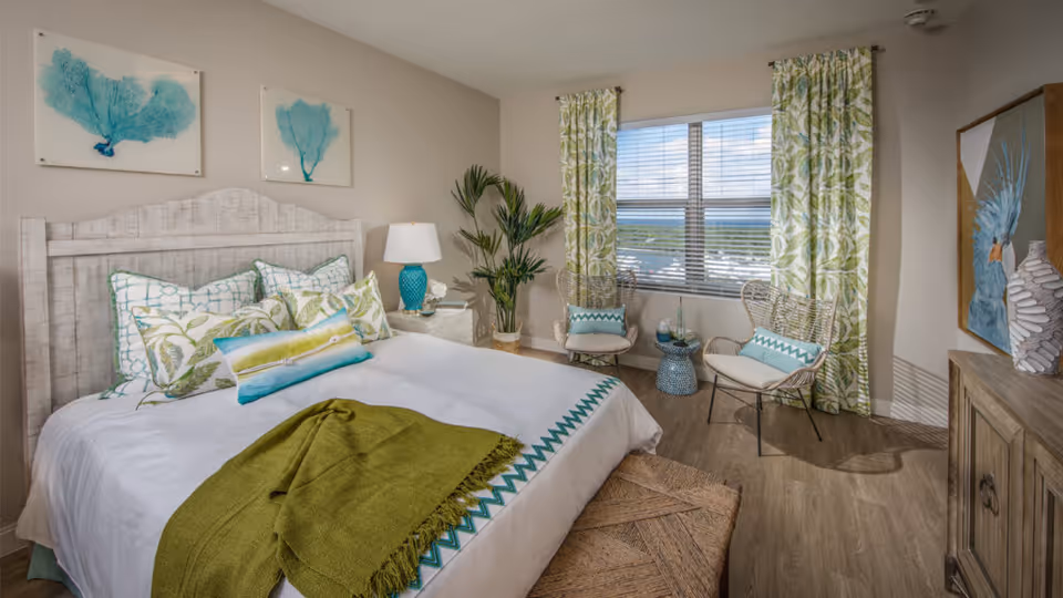 Bright coastal-themed bedroom with a whitewashed wooden bed, patterned pillows and green throw, two rattan chairs by a window with a water view, and wall artwork.