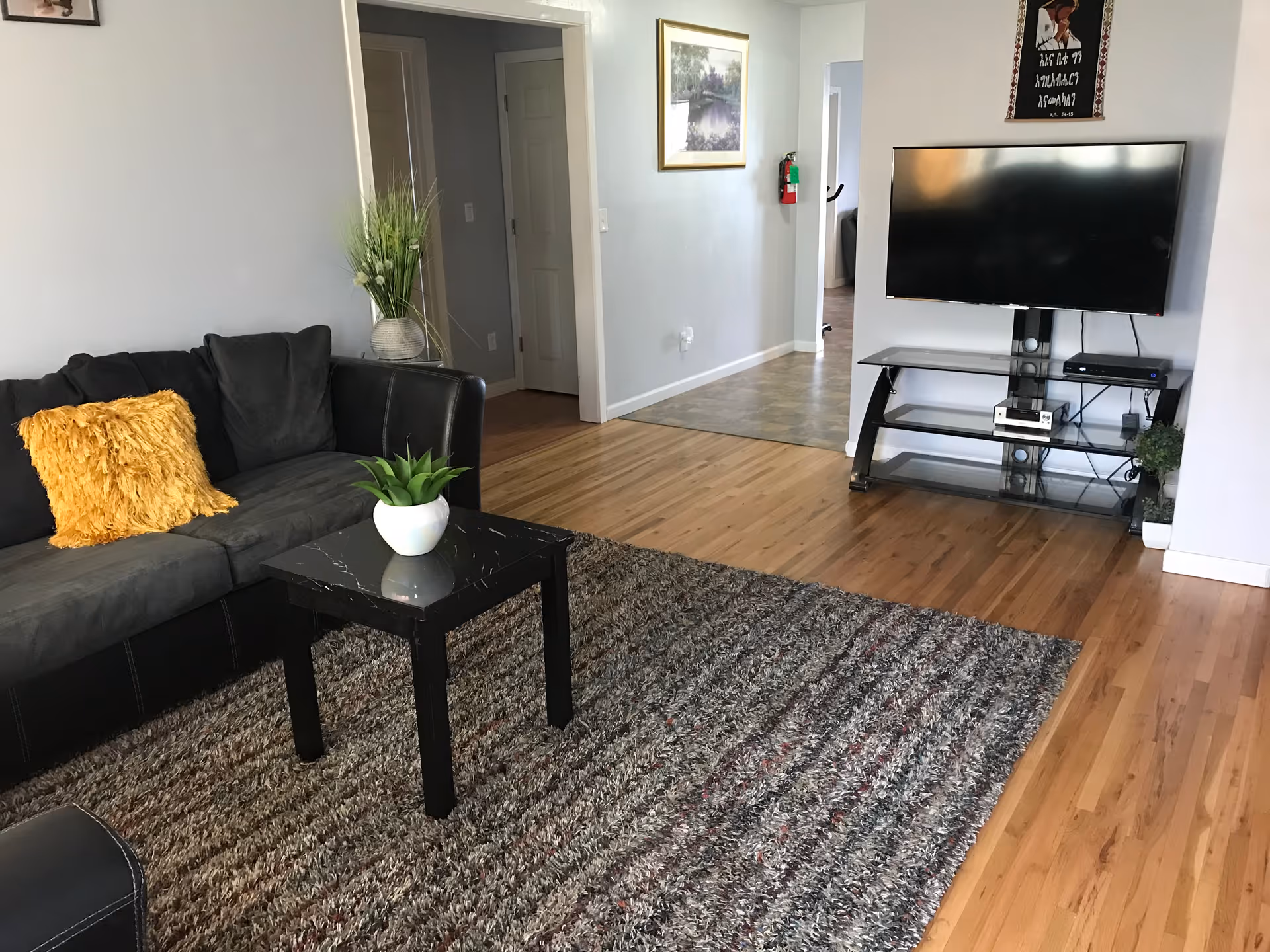A living room with a black and gray sectional sofa featuring a yellow throw pillow, a small black coffee table with a white potted plant, a large textured area rug, and a flat-screen TV mounted on a black TV stand. The room has light wood flooring and light gray walls with framed artwork and a fire extinguisher visible in the hallway.