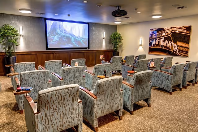 A cozy theater room with multiple rows of patterned armchairs facing a large screen displaying a forest scene. The room has soft lighting, wood paneling on the walls, potted plants in the corners, and a large framed photo of a theater marquee on the right wall.