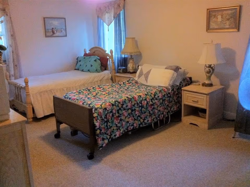 A cozy bedroom with two single beds, one with a floral bedspread and the other with a plain white bedspread. There are two nightstands, each with a lamp, and framed pictures hanging on the walls. A window with curtains allows natural light into the room.