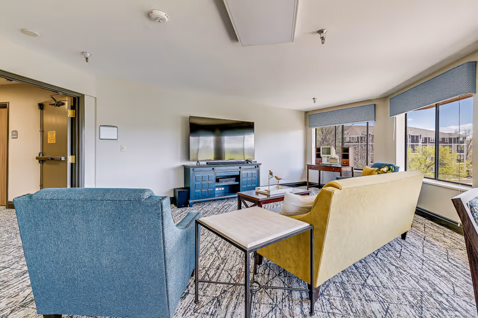 A bright living room in a senior living facility featuring a blue armchair and a yellow sofa facing a flat-screen TV mounted on a blue cabinet. The room has large windows with blue valances, allowing natural light to fill the space. A wooden side table and a coffee table with decorative items are also visible. Outside the windows, other buildings and trees can be seen.