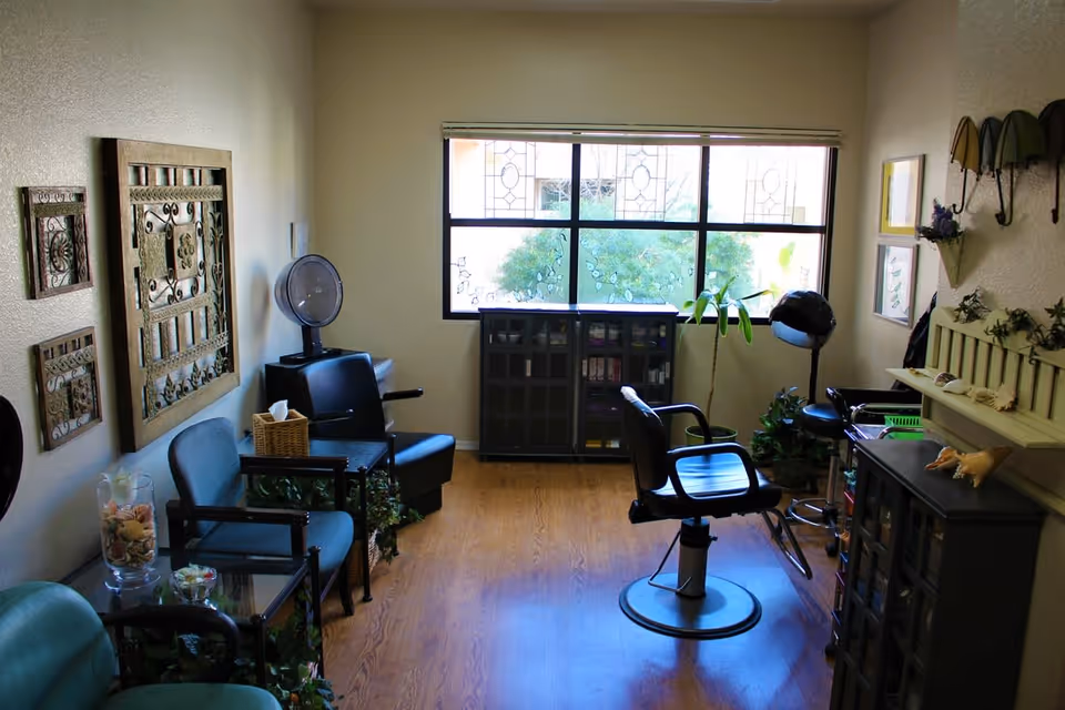 Interior of a small salon room with a salon chair in the center, additional seating along the left wall, decorative wall art, a large window with stained glass details, and various plants and salon equipment.