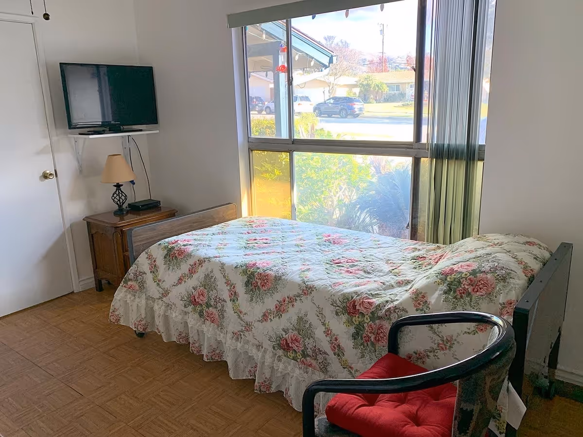 A small bedroom with a single bed covered in a floral quilt. Next to the bed is a wooden nightstand with a lamp and a device on it. A flat-screen TV is mounted on a white shelf on the wall above the nightstand. There is a large window with vertical blinds letting in natural light, and a chair with a red cushion is positioned near the foot of the bed. The floor has a wood-patterned surface.