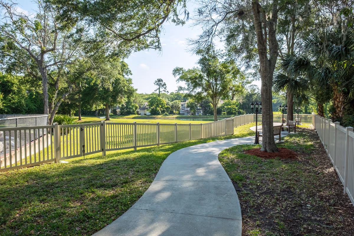 A winding concrete pathway bordered by a white fence runs through a green outdoor area with trees and benches. The scene is sunny with a clear sky and a calm, natural setting.