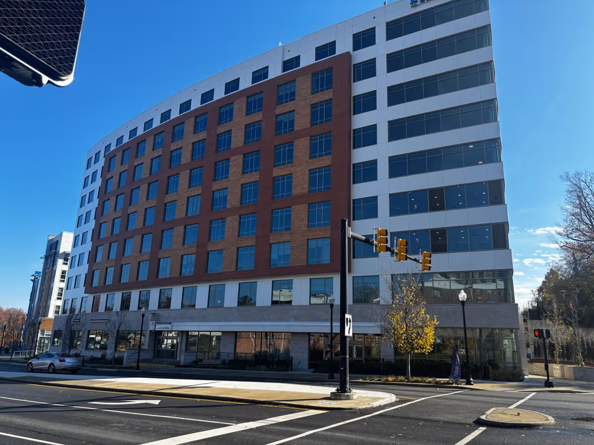 Street-level view of a multi-story modern brick-and-glass building at a city intersection under a clear blue sky.