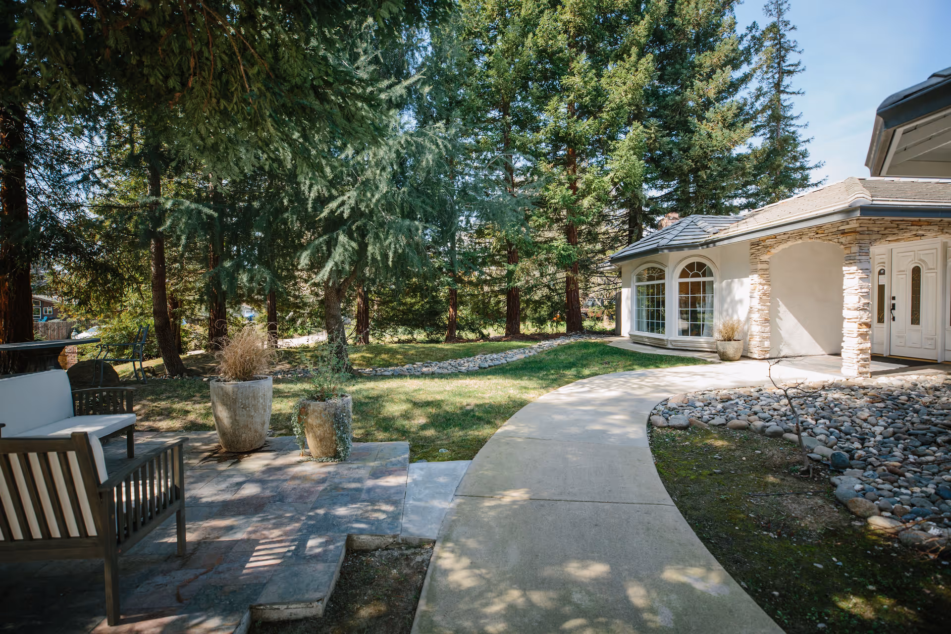Outdoor patio area with seating and potted plants next to a curved concrete walkway leading to a building entrance surrounded by trees and greenery.
