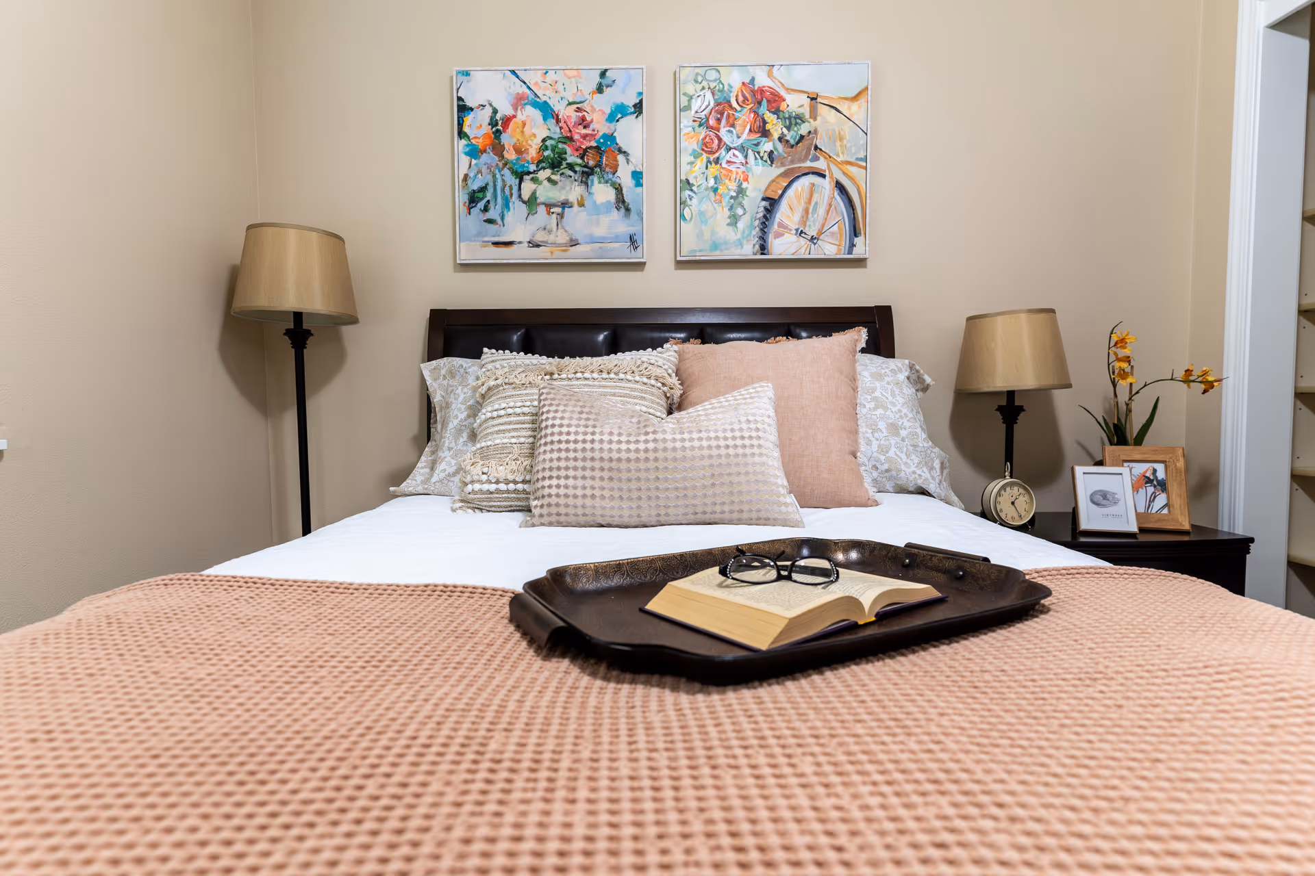 A neatly made bed with multiple decorative pillows and a textured pink blanket. On the bed is a dark tray holding an open book and a pair of glasses. On either side of the bed are matching nightstands with beige lamps. The right nightstand also has a small clock, a framed photo, and a vase with yellow flowers. Above the bed are two colorful floral paintings on a beige wall.