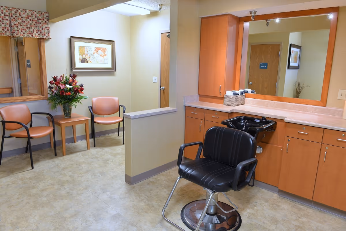 Interior view of a salon area in a senior living facility featuring a black salon chair in front of a large mirror and wooden cabinets with a black sink. To the left, there are two peach-colored chairs with black armrests next to a small wooden table holding a vase of flowers. A framed artwork hangs on the wall above the chairs.