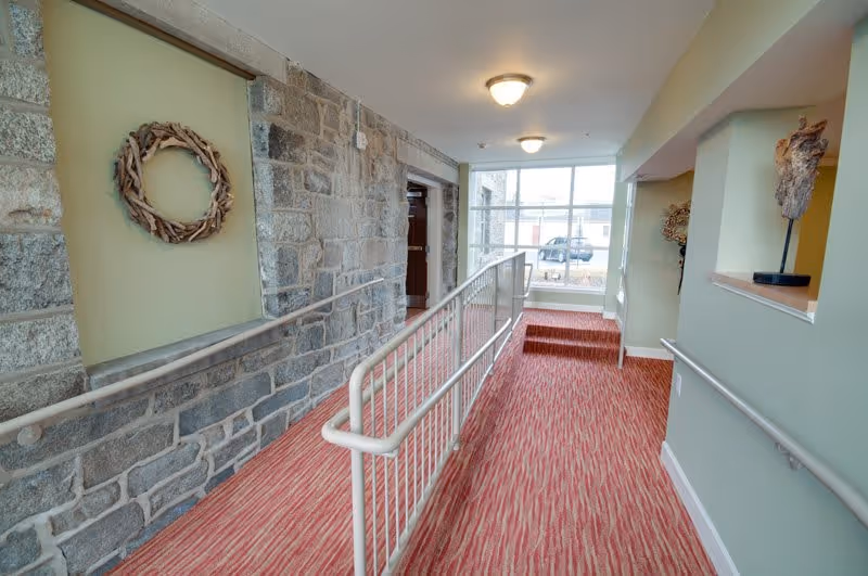 Interior hallway of a senior living facility with a red patterned carpet, a metal handrail ramp, stone wall on the left, light green painted walls, decorative wreath and sculpture, and a large window at the end showing a parked vehicle outside.