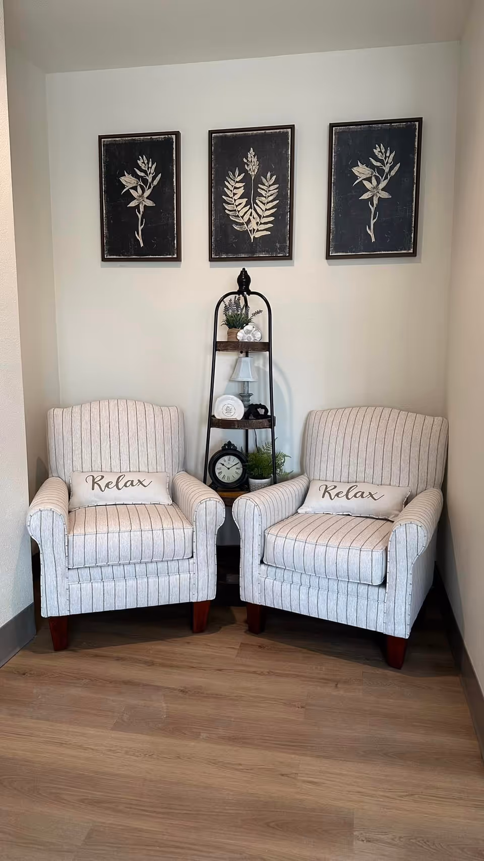 Two beige striped armchairs with 'Relax' pillows are placed side by side in a corner. Between them is a small black metal shelf unit holding decorative items including a clock, a small lamp, and plants. Above the chairs are three framed botanical prints on a light-colored wall. The floor is wooden.