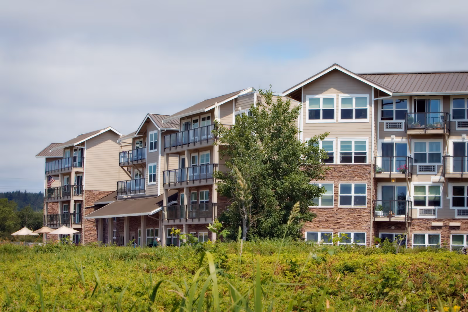 Multi-story senior living building with balconies and green landscaping in front under a cloudy sky.