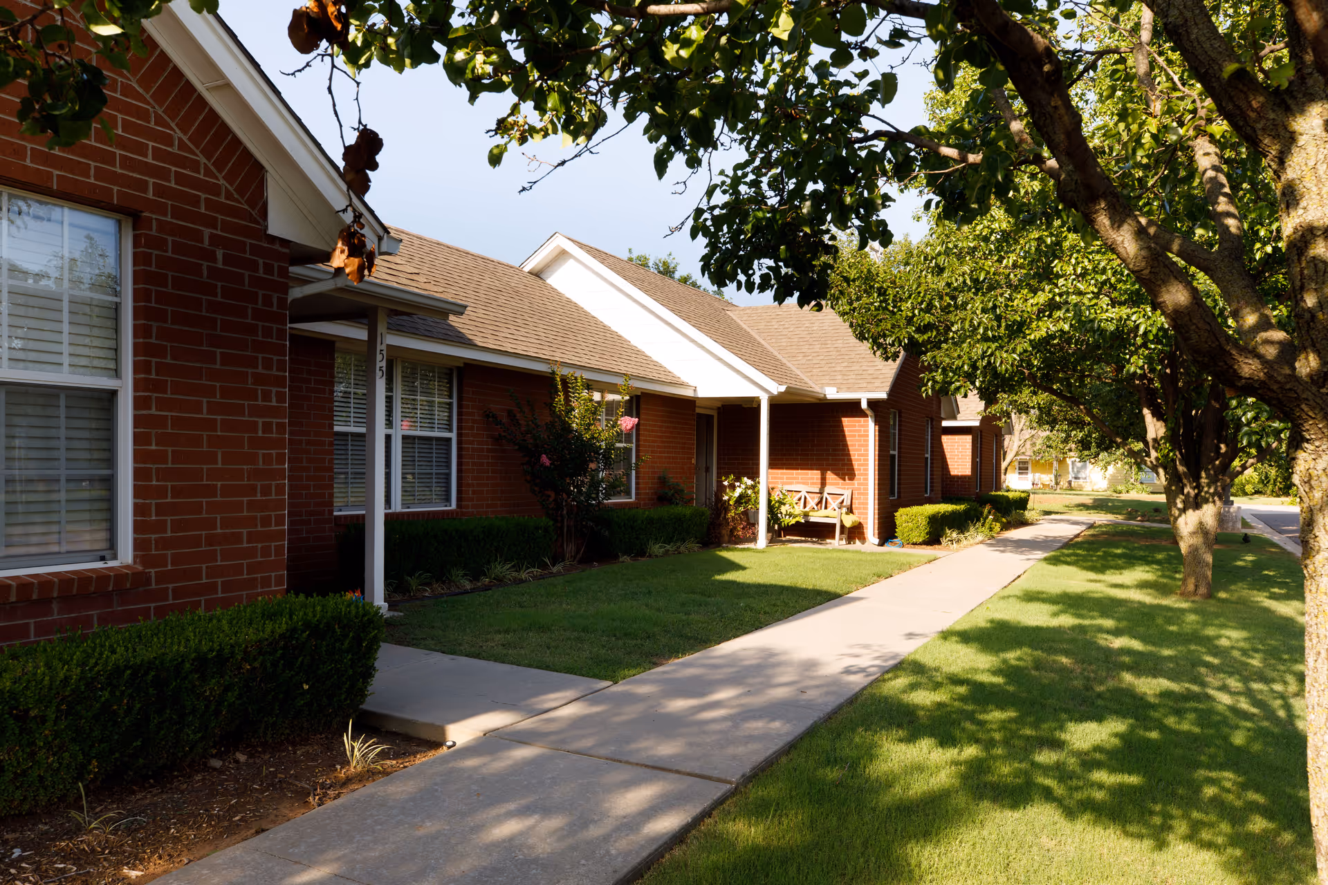 A sunny exterior view of a single-story brick building with a sidewalk leading to a covered entrance. The building is surrounded by well-maintained green grass, bushes, and trees casting shadows on the ground. There is a bench near the entrance under the covered porch.