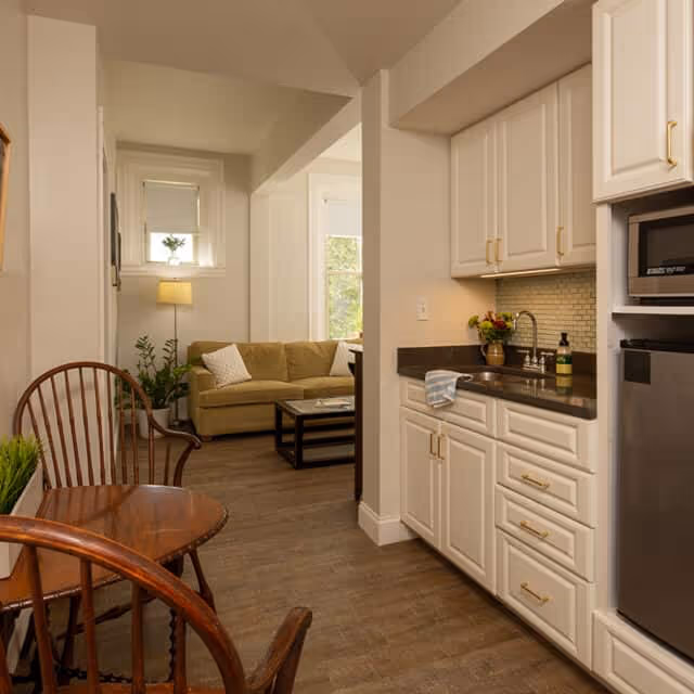 Interior view of a senior living facility showing a small kitchen area with white cabinets, a sink, a microwave, and a mini refrigerator. Adjacent to the kitchen is a cozy living room with a beige sofa, a coffee table, a floor lamp, and some plants near a window.
