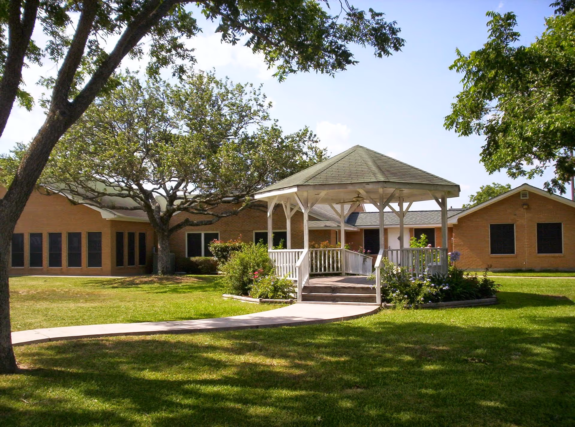 Outdoor view of a nursing and rehabilitation facility with a white gazebo in the center surrounded by green grass, trees, and bushes. The building is made of brick with multiple windows and a clear blue sky in the background.
