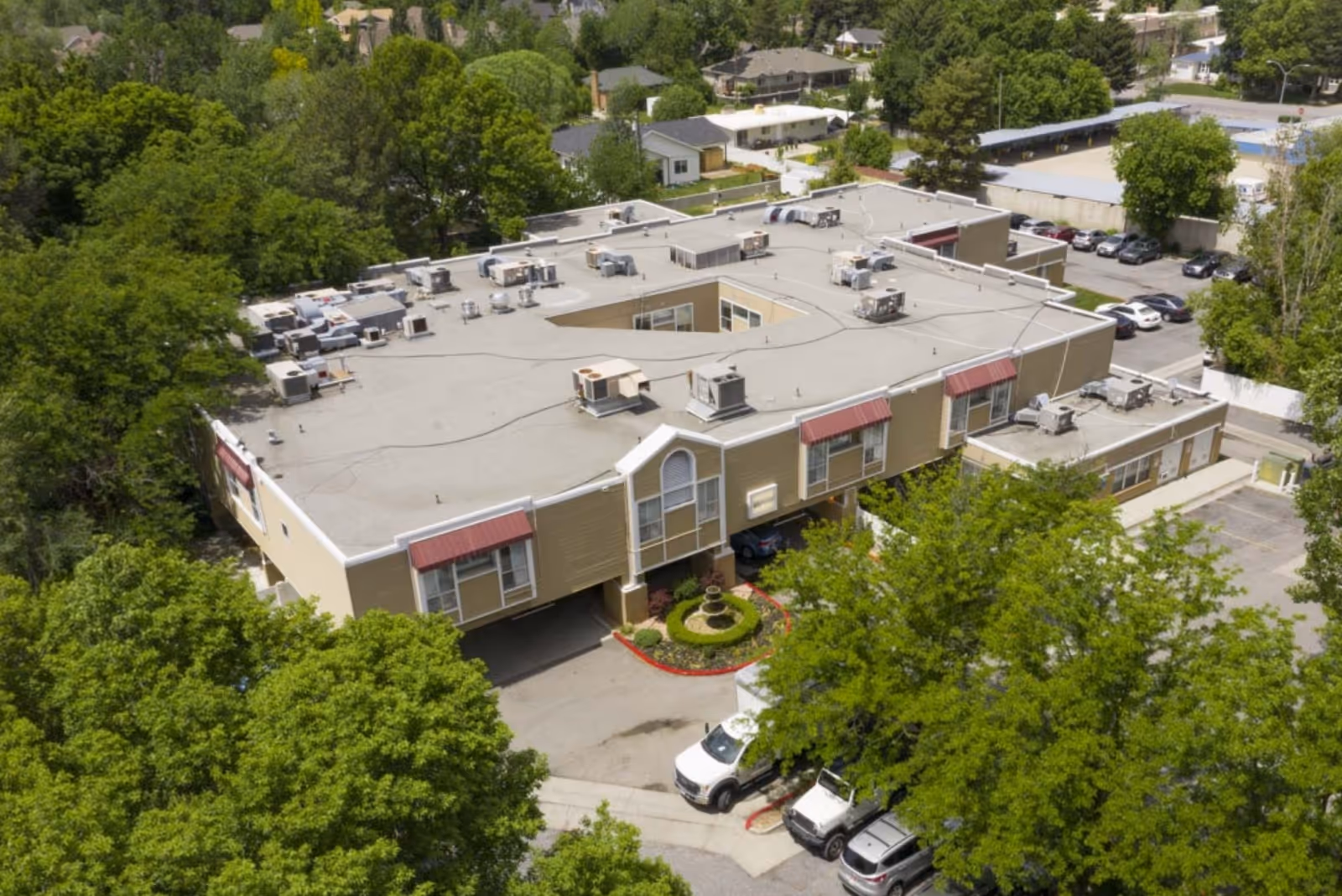Aerial view of a large, single-story healthcare facility building surrounded by trees and residential houses. The building has a flat roof with multiple HVAC units and a small courtyard in the center. Several cars are parked in the parking lot adjacent to the building.