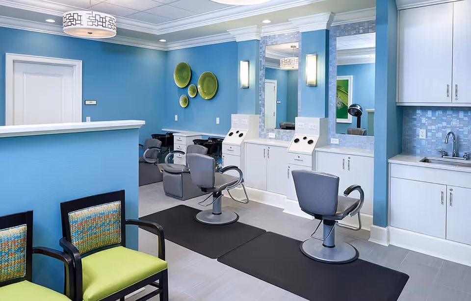 Interior view of a salon area in an assisted living facility with blue walls, two salon chairs in front of large mirrors, a small sink area with cabinets, and two green and patterned waiting chairs. The space is well-lit with ceiling and wall lights, and decorative green plates are mounted on the wall.
