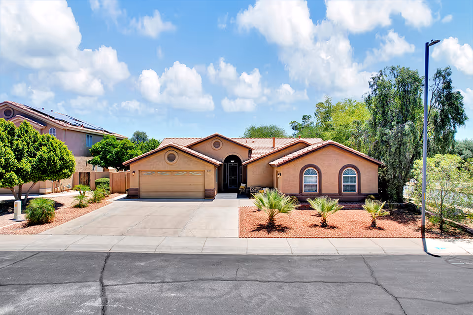 Front exterior view of a single-story residential building with a two-car garage, beige stucco walls, and a red tile roof. The driveway and front yard are landscaped with small palm trees and bushes, under a partly cloudy blue sky.