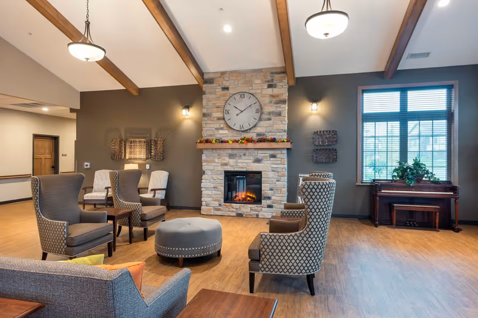A cozy living room area with wooden beams on the ceiling, a stone fireplace with a large clock above it, several upholstered armchairs arranged around a round ottoman, a window with blinds, and a piano with a plant on top.