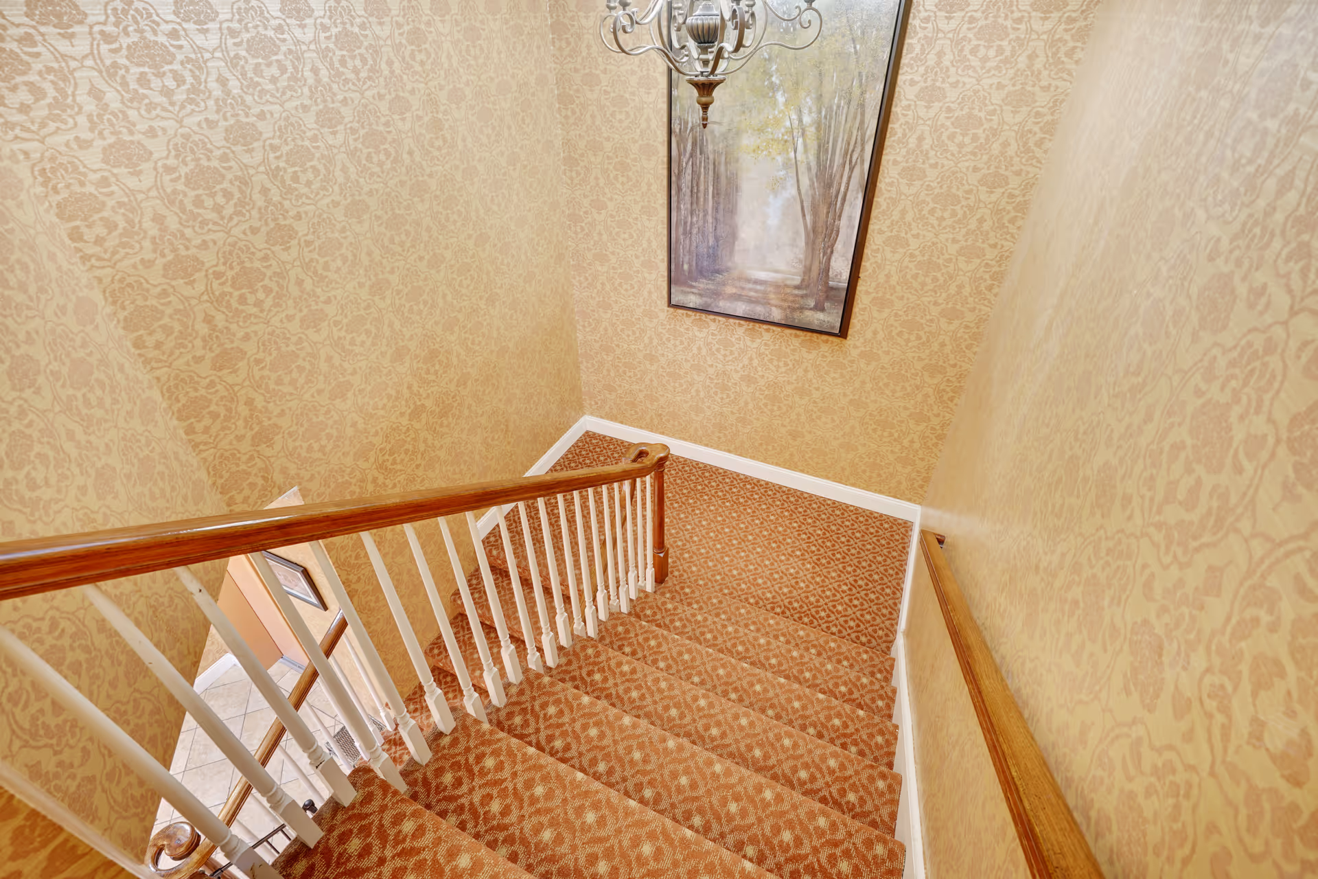 View looking down a carpeted staircase with a wooden handrail and white balusters. The walls are covered with patterned wallpaper in beige tones, and a framed painting of a forest path hangs on the wall at the bottom of the stairs. A decorative chandelier is visible above the stairs.