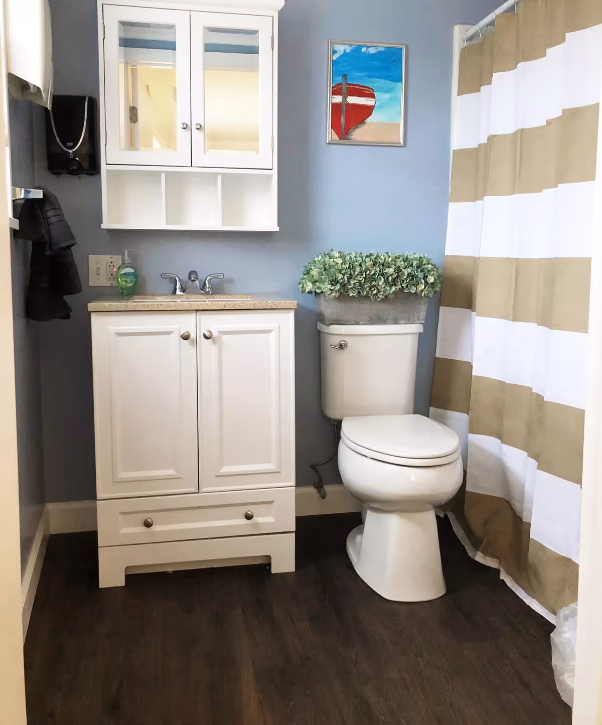 A small bathroom with a white vanity and mirrored cabinet, a toilet topped with a potted plant, and a beige-and-white striped shower curtain.